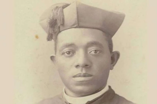 A man in a clerical outfit and cap looks directly at the camera. The image has a sepia tone, with a plain background, conveying a formal mood.
