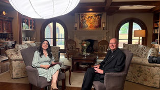 Two people sit in armchairs in an elegant living room with a fireplace, sofas, and a large white overhead light, smiling.