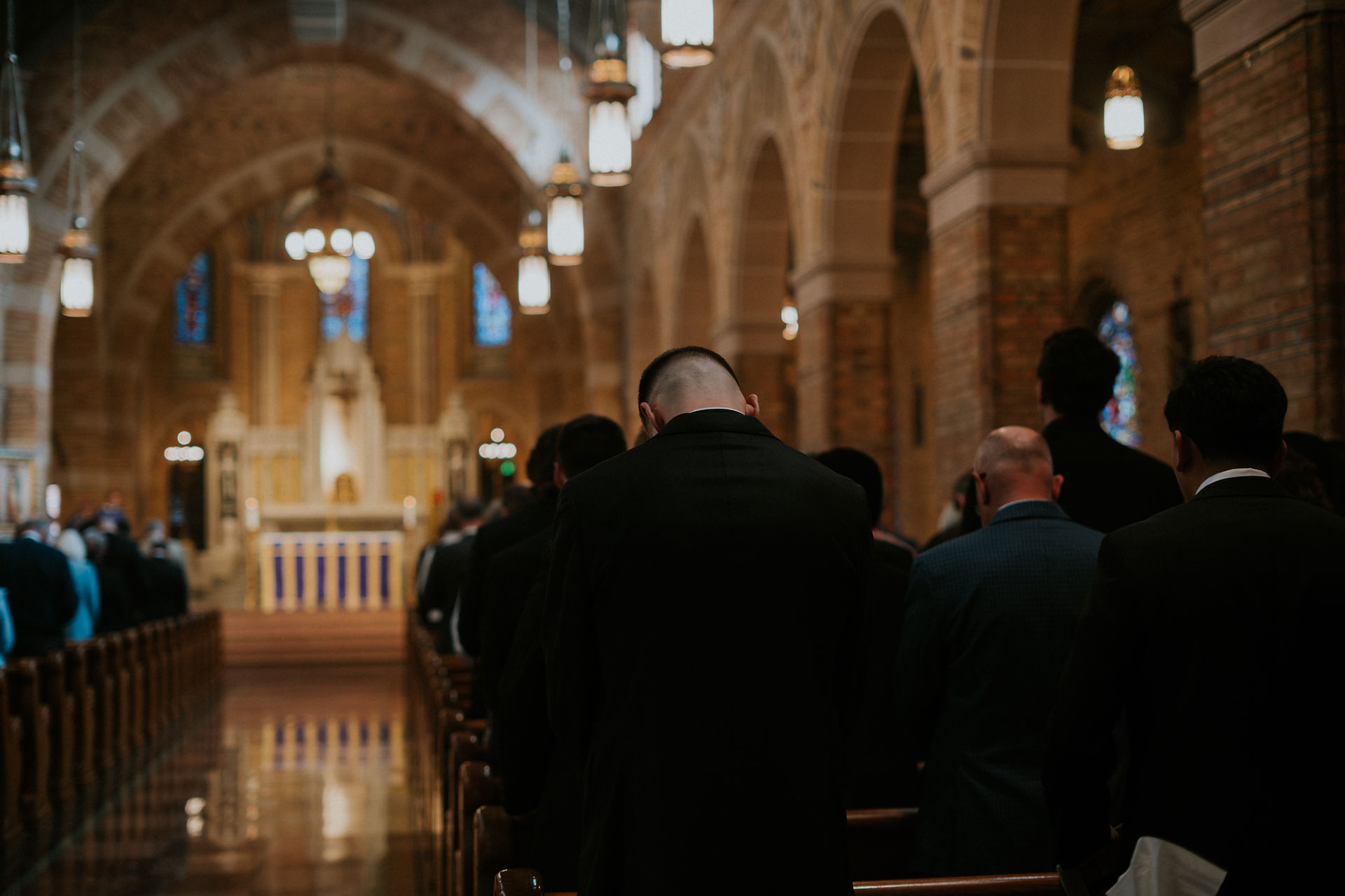 People in suits seated in a dimly lit church, facing an altar with stained glass windows. Mood is solemn and introspective.