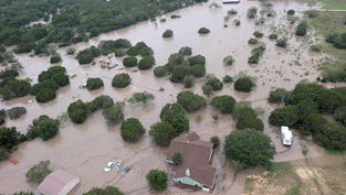 Aerial view of flooded rural area with homes and vehicles partially submerged. Brown floodwater surrounds green trees, creating a somber mood.