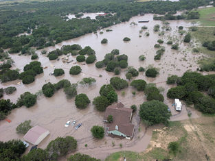 Aerial view of flooded rural area with homes and vehicles partially submerged. Brown floodwater surrounds green trees, creating a somber mood.