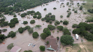 Aerial view of flooded rural area with homes and vehicles partially submerged. Brown floodwater surrounds green trees, creating a somber mood.