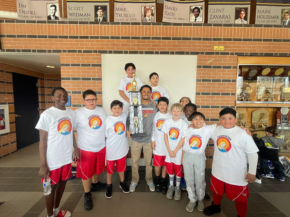 Group of kids and a coach smiling with a large trophy indoors. They're wearing white Annunciation shirts and red shorts. Brick background.