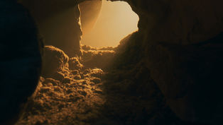 Sunlight streams into a rocky cave entrance, casting warm orange and brown hues on the sandy floor, creating a serene atmosphere.