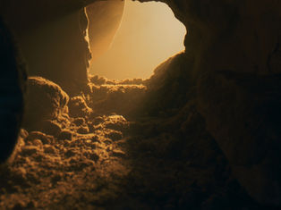 Sunlight streams into a rocky cave entrance, casting warm orange and brown hues on the sandy floor, creating a serene atmosphere.