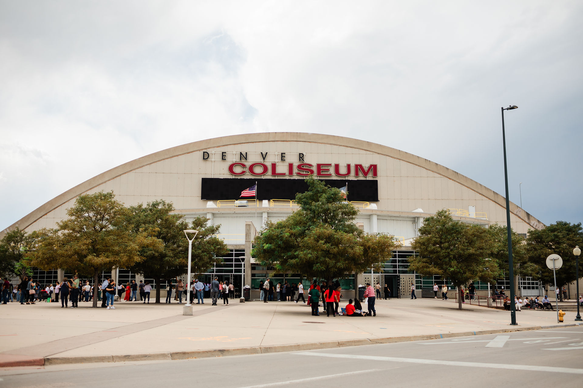 People gather outside the Denver Coliseum, a large dome-shaped building. Trees and a few American flags are visible. The sky is overcast.