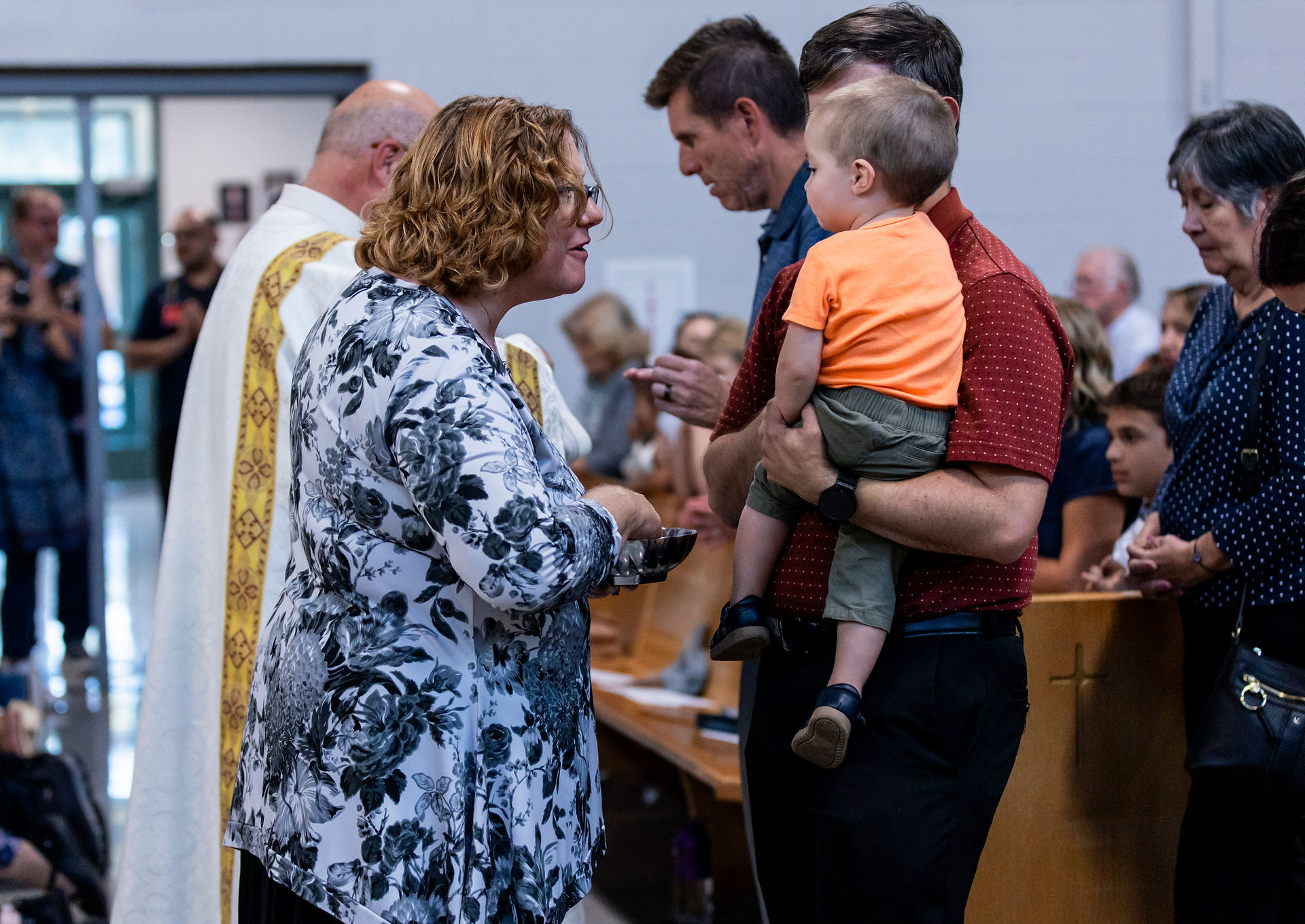 Woman in floral shirt giving communion to man holding a child in church. Congregation seated in background. Calm atmosphere.