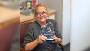 Smiling person holding a clear award plaque in an office. Background shows a kitchen area. Wearing glasses and a grey shirt.