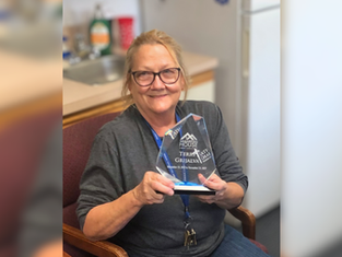 Smiling person holding a clear award plaque in an office. Background shows a kitchen area. Wearing glasses and a grey shirt.
