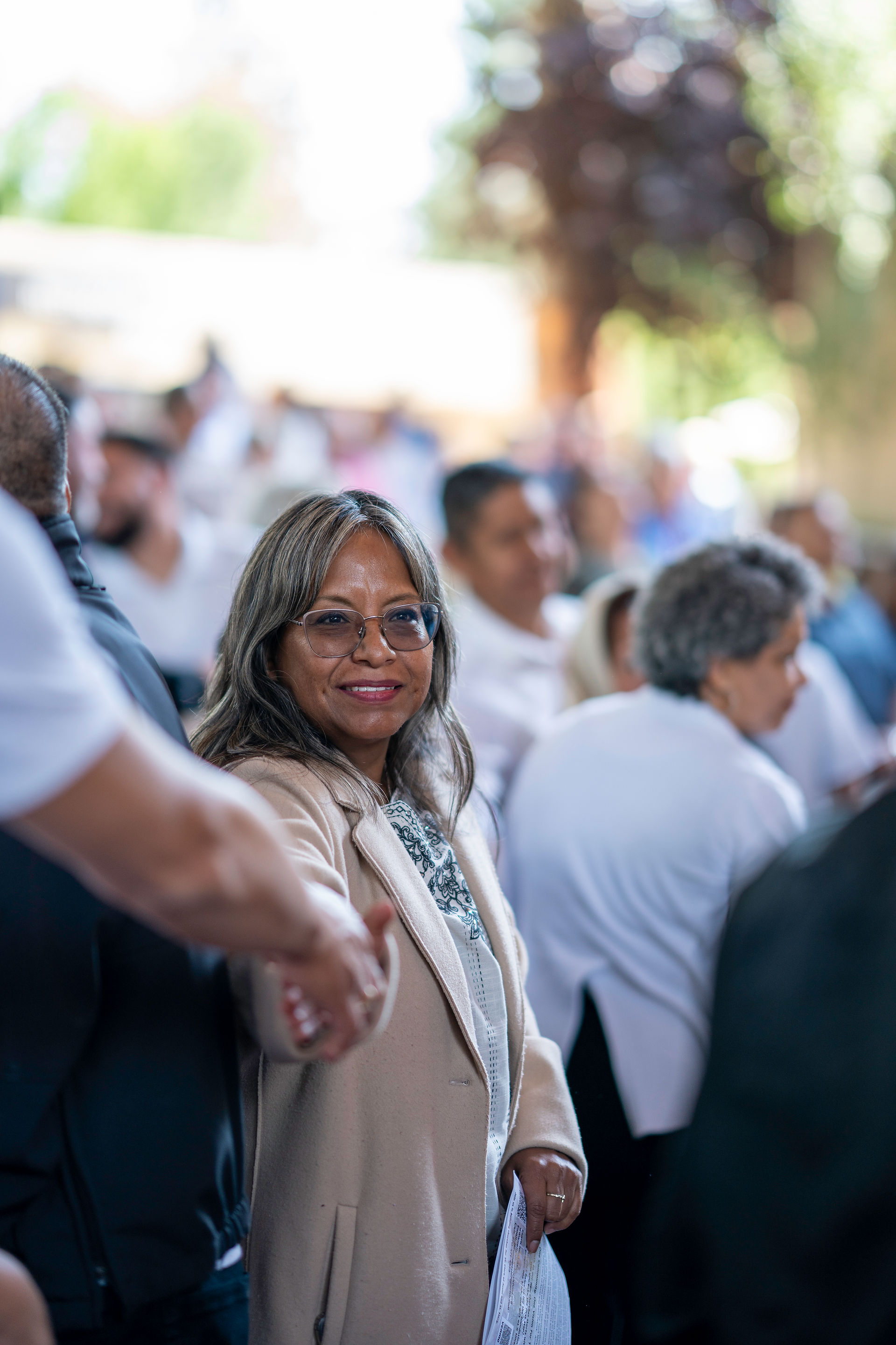 Smiling woman in glasses shakes hands in a crowd, holding a paper. Background features blurred people in white, outdoors with greenery.