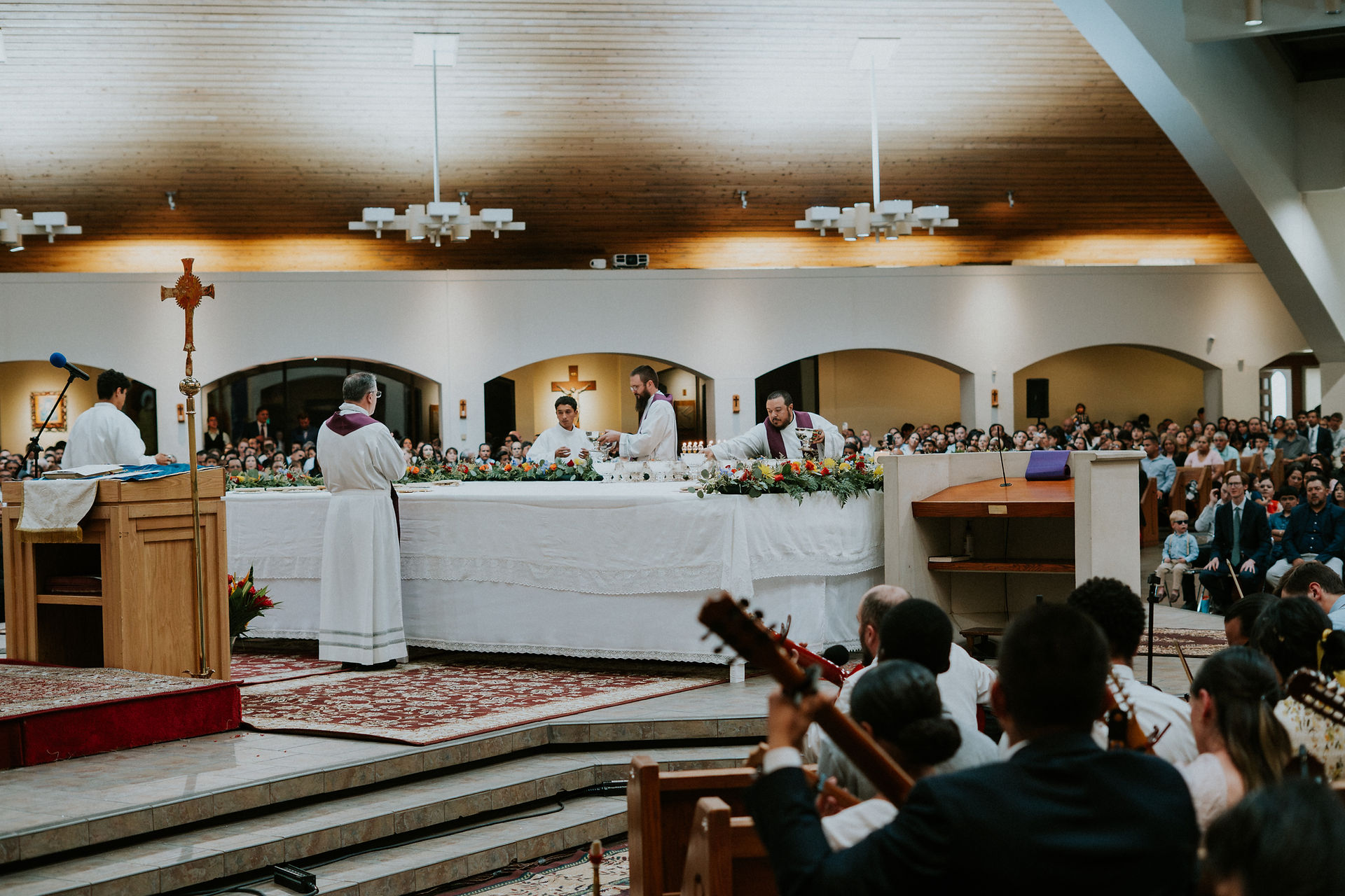 A church service with clergy at the altar, surrounded by a large congregation. The setting is bright with wooden ceilings and floral decor.