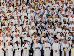 A large group of priests in white robes stands in rows, holding hands in a prayer gesture. The mood is solemn and reverent.