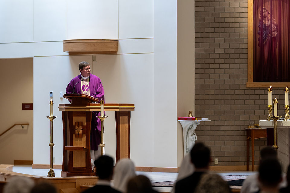 A priest in a purple robe speaks at a wooden lectern in a church. Candles and altar visible; audience blurred in foreground. Calm atmosphere.