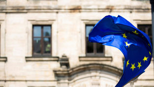 European Union flag with yellow stars waving in the wind in front of a historic stone building with arched windows.