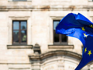 European Union flag with yellow stars waving in the wind in front of a historic stone building with arched windows.