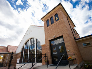 Brick and white church with arched windows against a blue sky, entrance labeled "925," surrounded by a black fence and potted flowers.