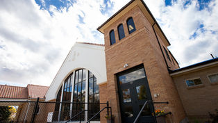 Brick and white church with arched windows against a blue sky, entrance labeled "925," surrounded by a black fence and potted flowers.