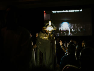 A person holds a large circular object in a dim room. People raise hands. Projected text reads "Your name stands above them all."