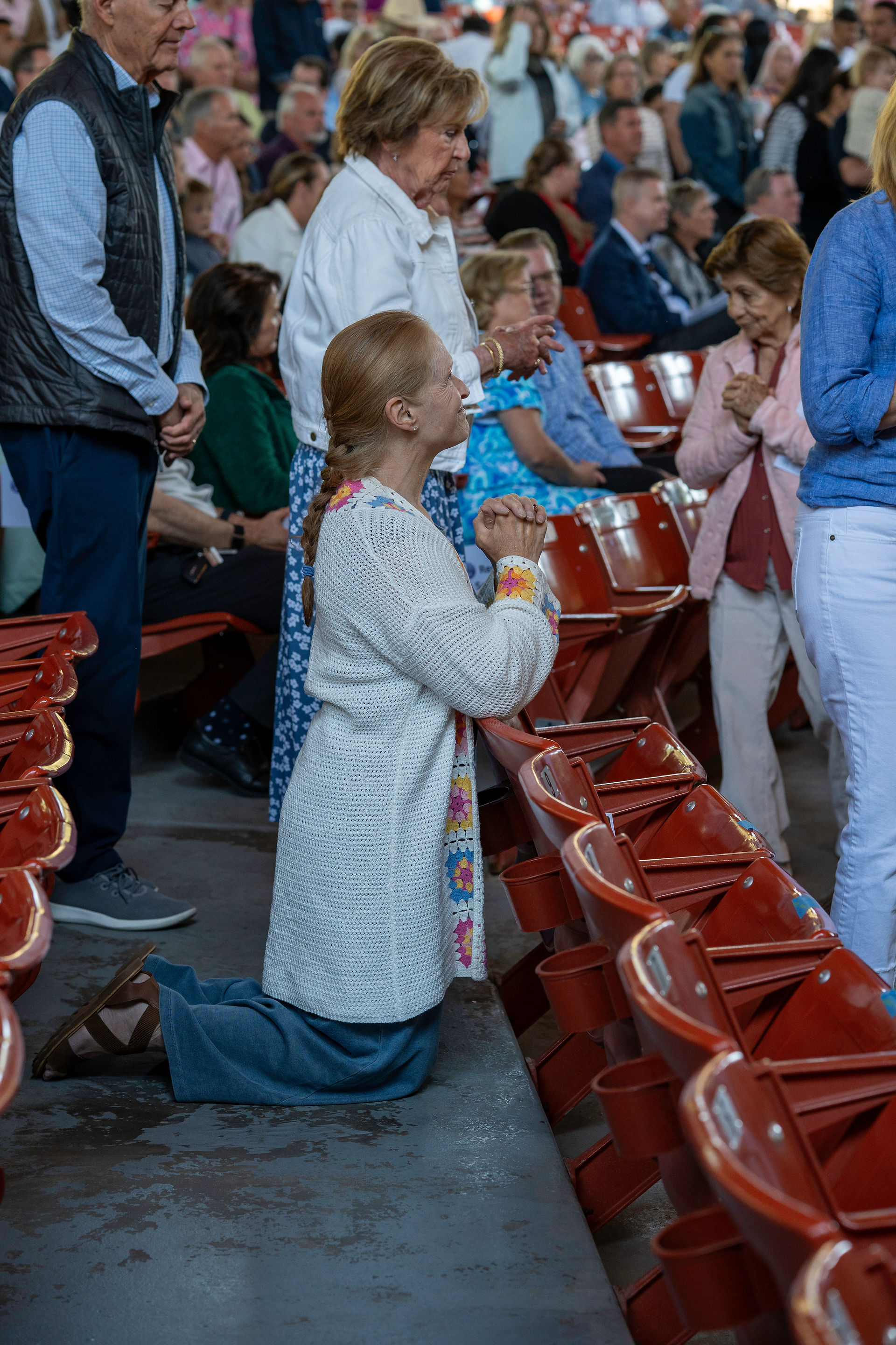 A group of people gathered in a religious setting, with one person kneeling between rows of red seats, hands clasped in prayer. Others are standing or seated nearby, also appearing to be in prayer or reflection.