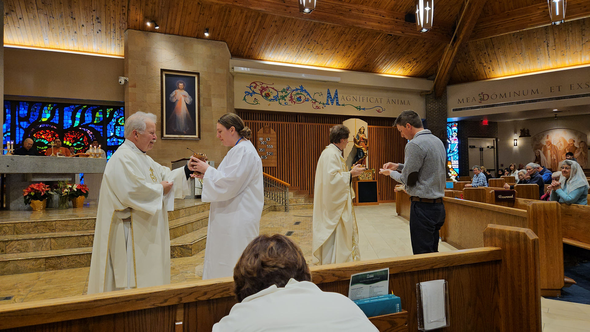 Clergy in white robes distribute communion in a church. Stained glass and religious art adorn the wooden interior. Congregation is seated.