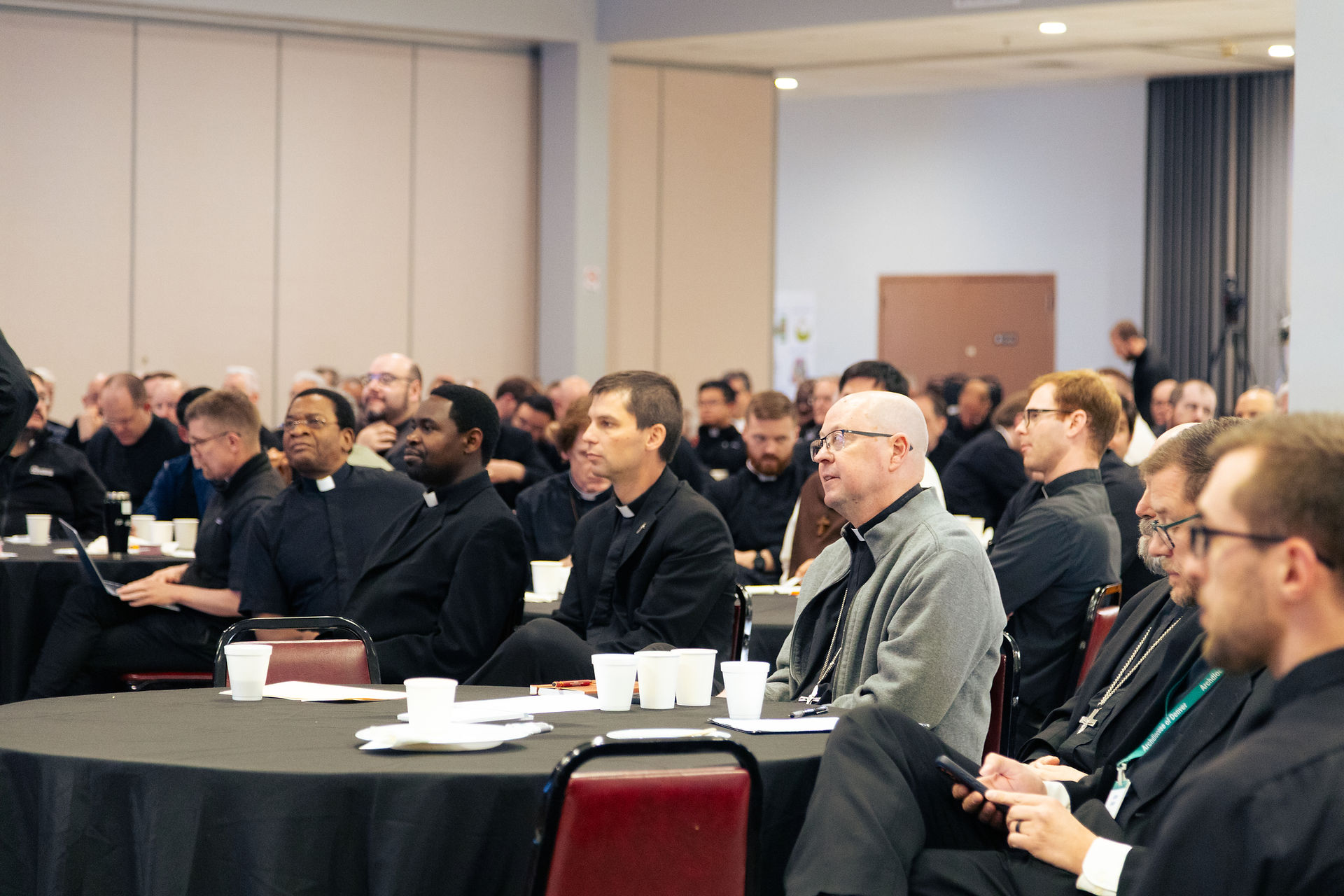 Group of Catholic clergy sit at round tables listening attentively during a meeting or conference.