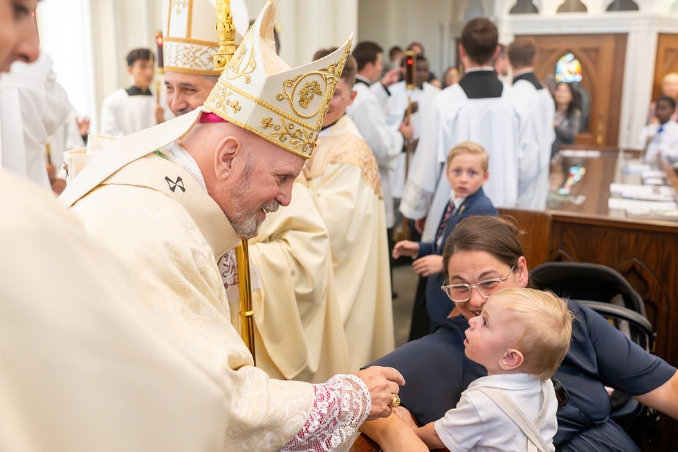 Bishop in gold vestments and mitre smiles and greets a young child held by his mother during a church liturgy, with clergy and altar servers in the background.