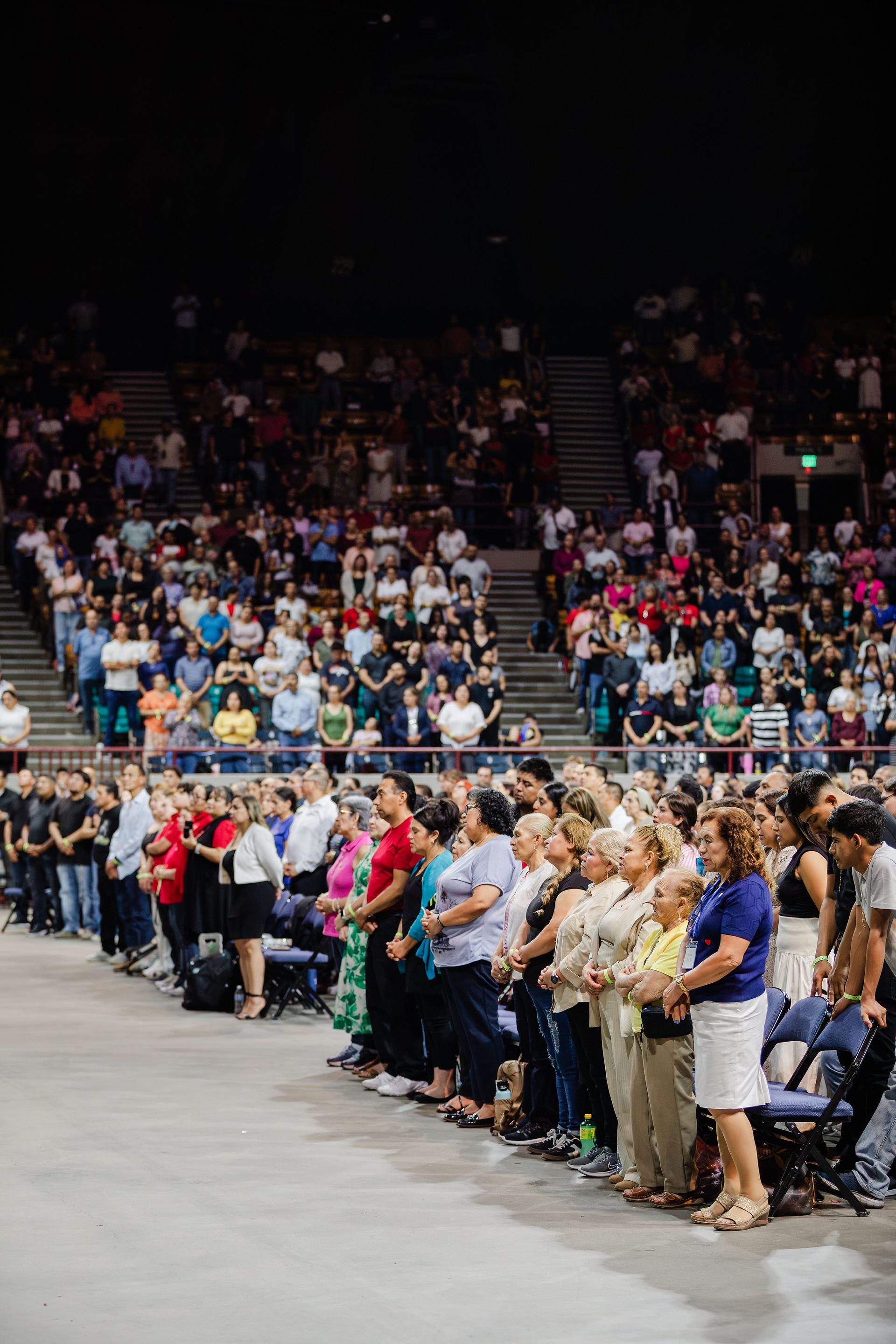 Crowd stands solemnly in a large auditorium. Rows of people in colorful attire face forward. Moods appear serious and attentive.