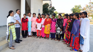Smiling women and a man stand in a semicircle outside a building, wearing colorful salwar kameez and chatting.