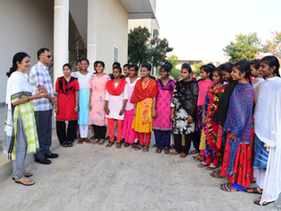 Smiling women and a man stand in a semicircle outside a building, wearing colorful salwar kameez and chatting.