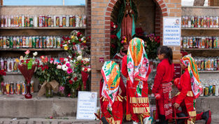 People in red outfits and colorful scarves kneel in front of a shrine with candles and flowers. A sign reads "AVOID THEFT PLEASE PLACE YOUR OFFERING."