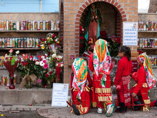 People in red outfits and colorful scarves kneel in front of a shrine with candles and flowers. A sign reads "AVOID THEFT PLEASE PLACE YOUR OFFERING."