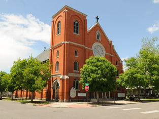 Red brick church with arched windows, cross on top, and trees in front. A stop sign is visible; it's a sunny day with a few clouds.