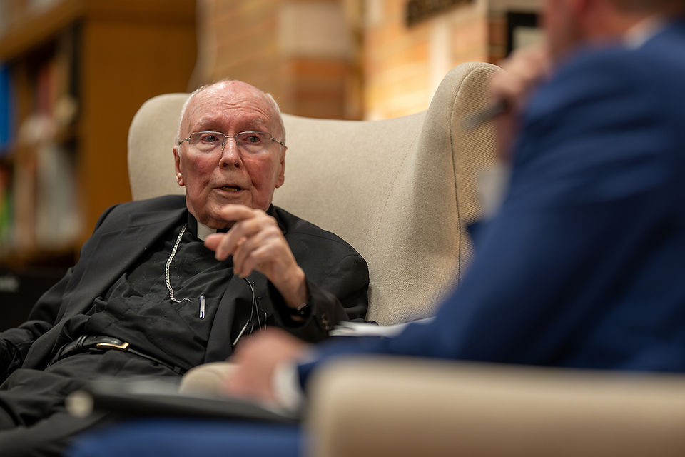 Elderly man in black clerical attire, gesturing while talking to a person in blue suit. They're seated in a cozy room with blurred bookshelves.