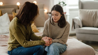 Two women sit on a bed holding hands in a cozy room with soft lighting. One wears a green cardigan, the other a beige sweater, both appear thoughtful.