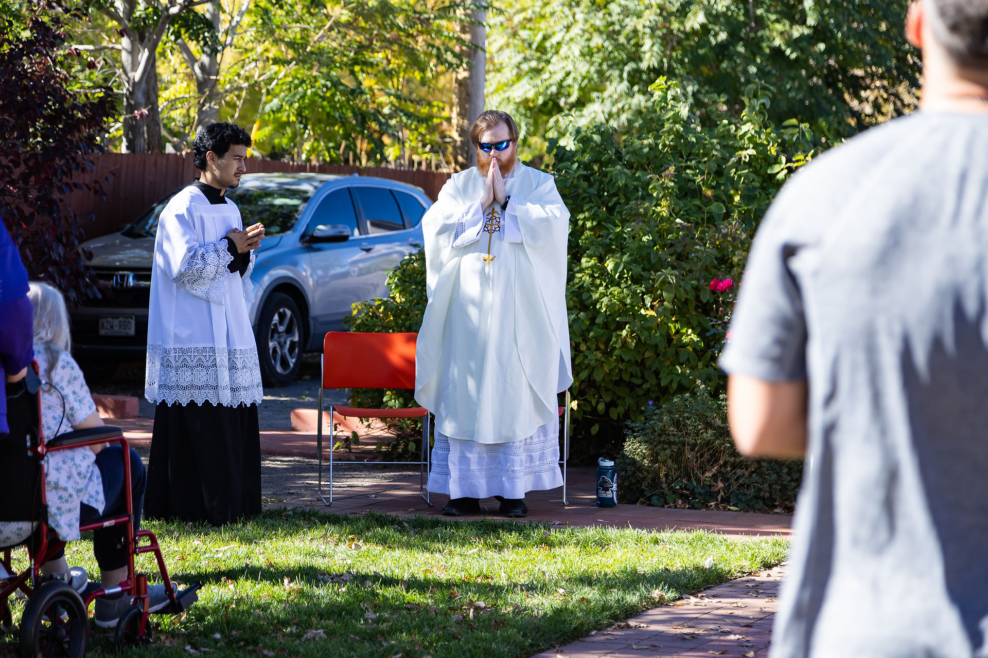 Priest and altar server in white robes praying outdoors with greenery in the background. Sunlit scene with a wheelchair and chairs visible.