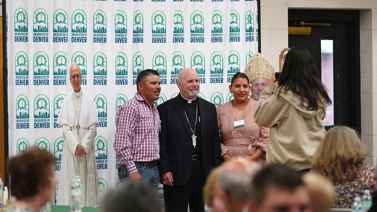 Three people pose for a photo in front of an "Archdiocese of Denver" backdrop. Another person photographs them. Cardboard cutouts visible.
