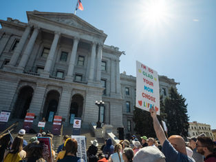 Protest outside a grand building under bright sun. Crowd holds signs; one reads "I'M SO GLAD YOUR MOM CHOSE LIFE." Energetic atmosphere.