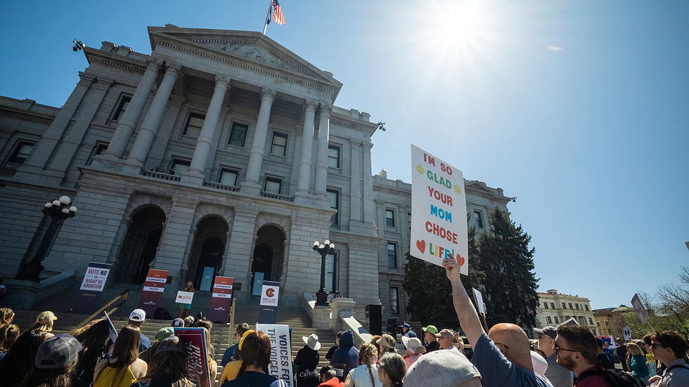 Protest outside a grand building under bright sun. Crowd holds signs; one reads "I'M SO GLAD YOUR MOM CHOSE LIFE." Energetic atmosphere.