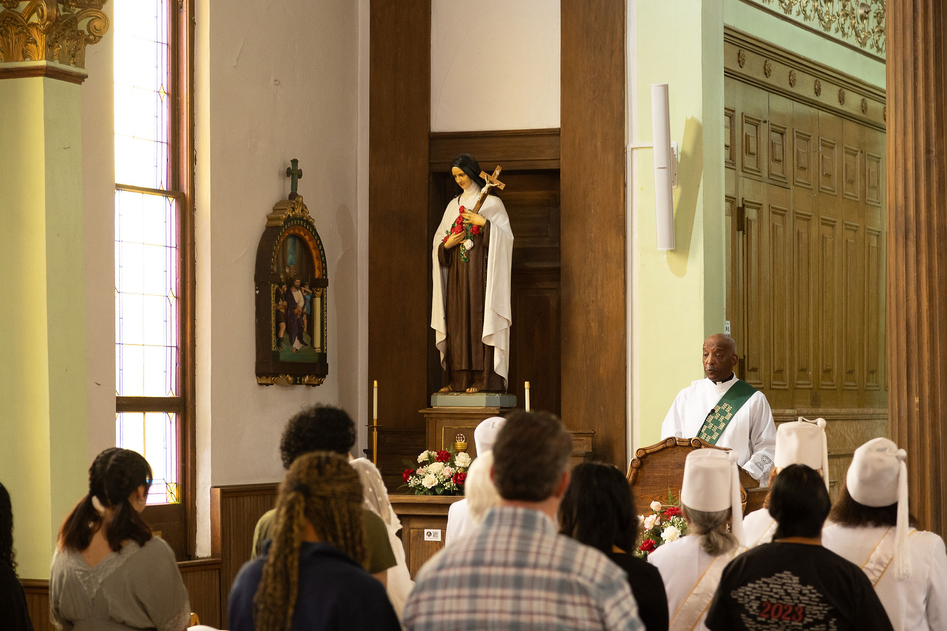 A deacon speaks to a standing congregation in a church with a stained glass window. A statue and floral arrangements are visible nearby.