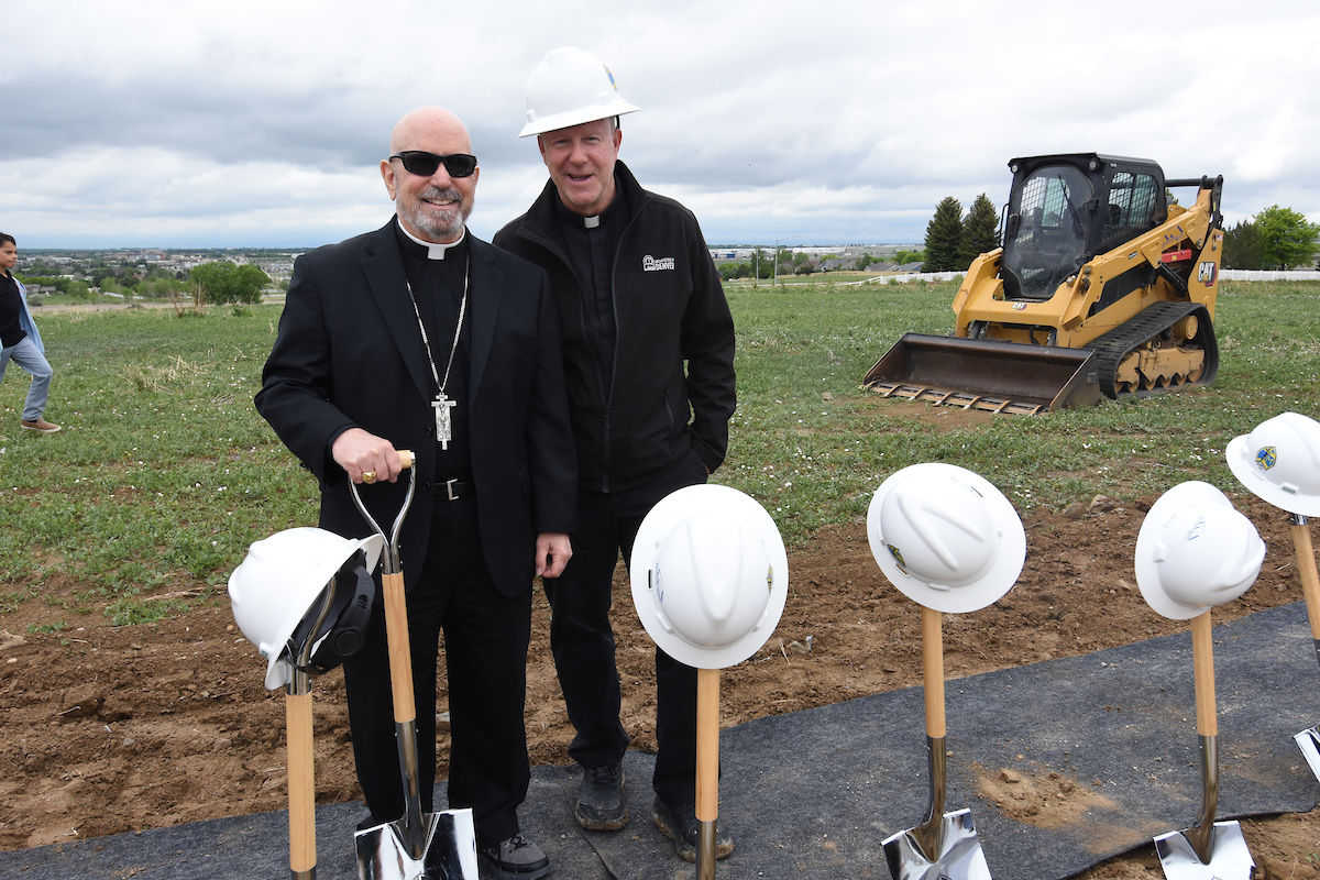Two men in black suits and hard hats pose with shovels at a construction site. A yellow bulldozer is in the grassy background.