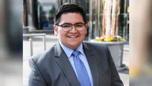 Smiling person in glasses wearing a gray suit and blue tie, standing outdoors with tall glass buildings and potted flowers in the background.