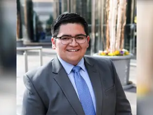 Smiling person in glasses wearing a gray suit and blue tie, standing outdoors with tall glass buildings and potted flowers in the background.