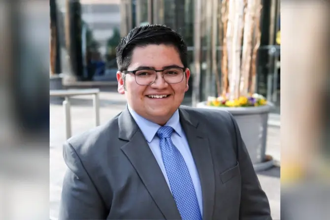 Smiling person in glasses wearing a gray suit and blue tie, standing outdoors with tall glass buildings and potted flowers in the background.