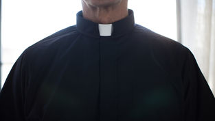 Close-up of a person wearing a black clerical shirt with a white collar. Bright light illuminates the background, creating a serene mood.
