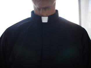 Close-up of a person wearing a black clerical shirt with a white collar. Bright light illuminates the background, creating a serene mood.