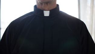 Close-up of a person wearing a black clerical shirt with a white collar. Bright light illuminates the background, creating a serene mood.