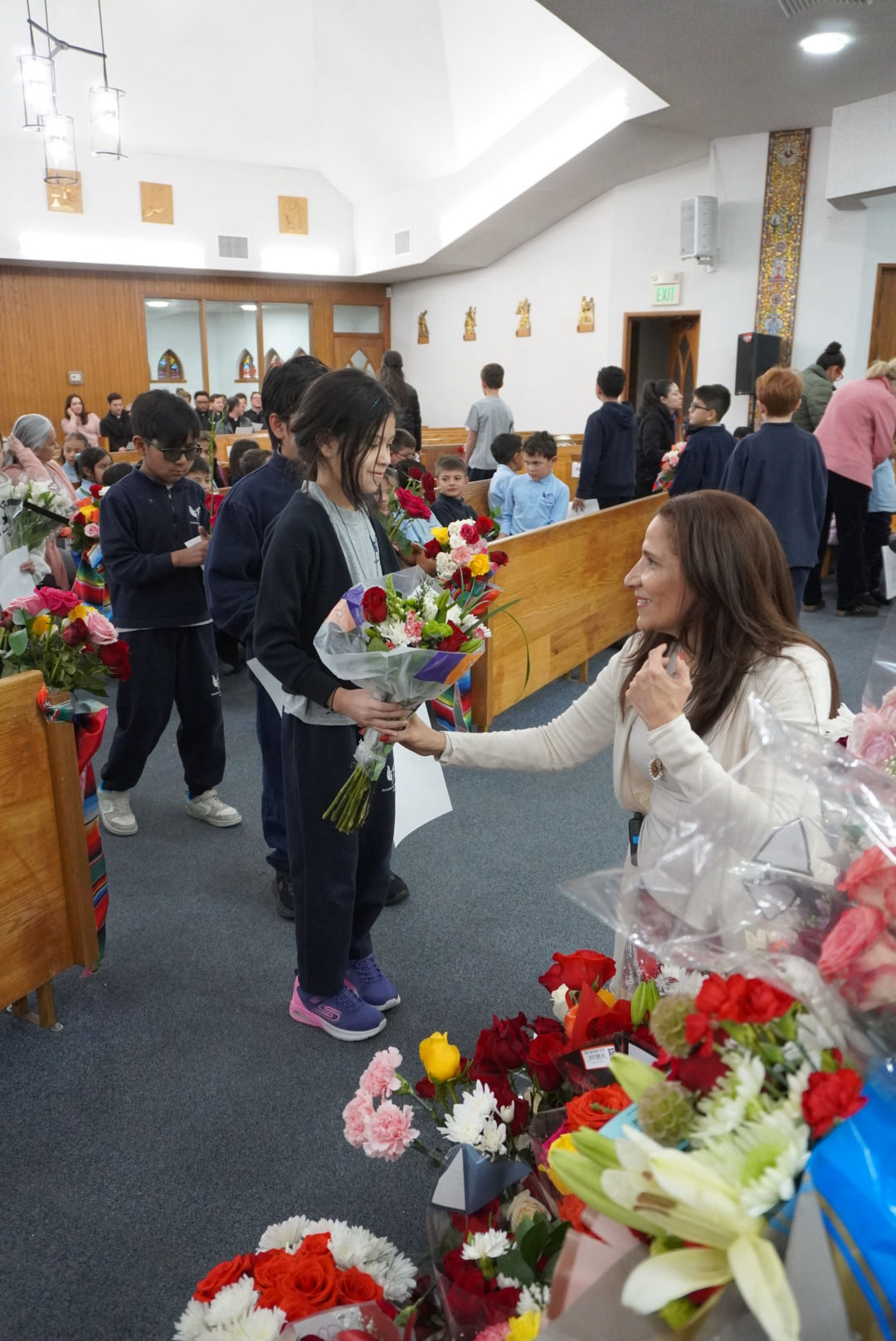 Girl with bouquet smiling at woman in church setting. People in background, colorful flowers around, creating a joyful atmosphere.