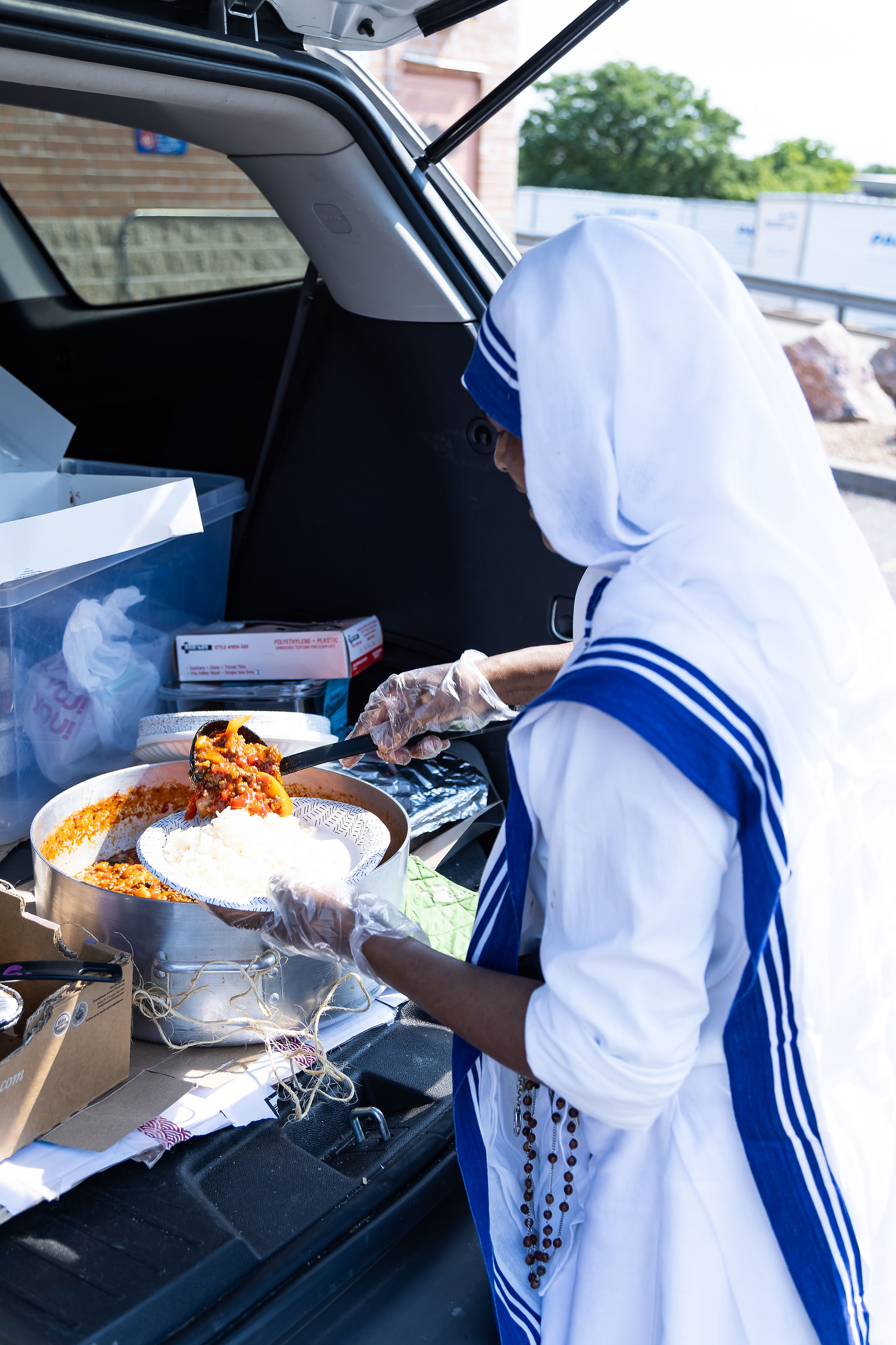 Woman in white and blue clothing serving food from a pot into a plate from open trunk. Boxes and containers visible. Outdoor scene.