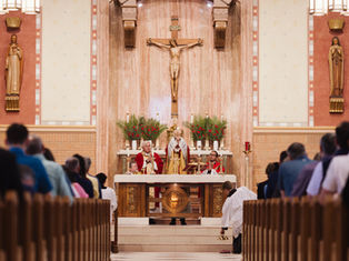 Catholic mass in a church: priests at the altar beneath a crucifix, with congregation kneeling in the pews.
