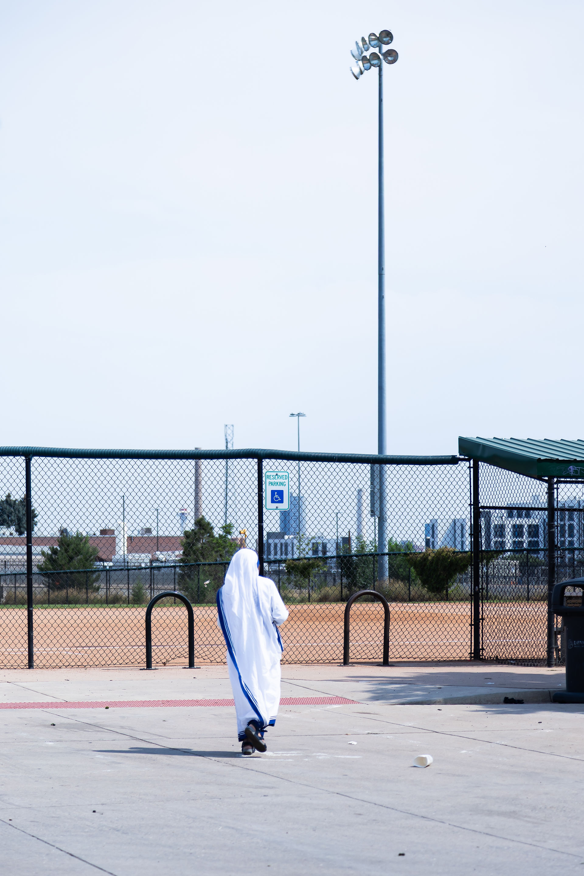 Person in a religious habit walking on concrete, near a fence with "Reserved Parking" sign. Bright day, industrial background.